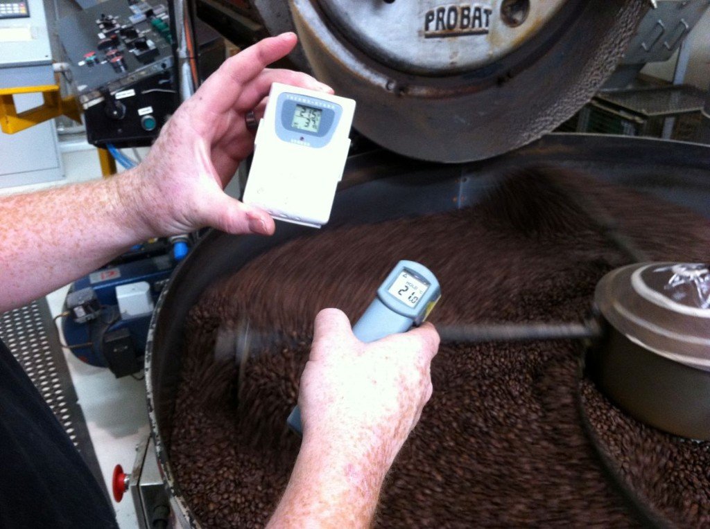 Infrared thermometer aimed at roasted coffee beans in a cooling tray beside a temperature and humidity display