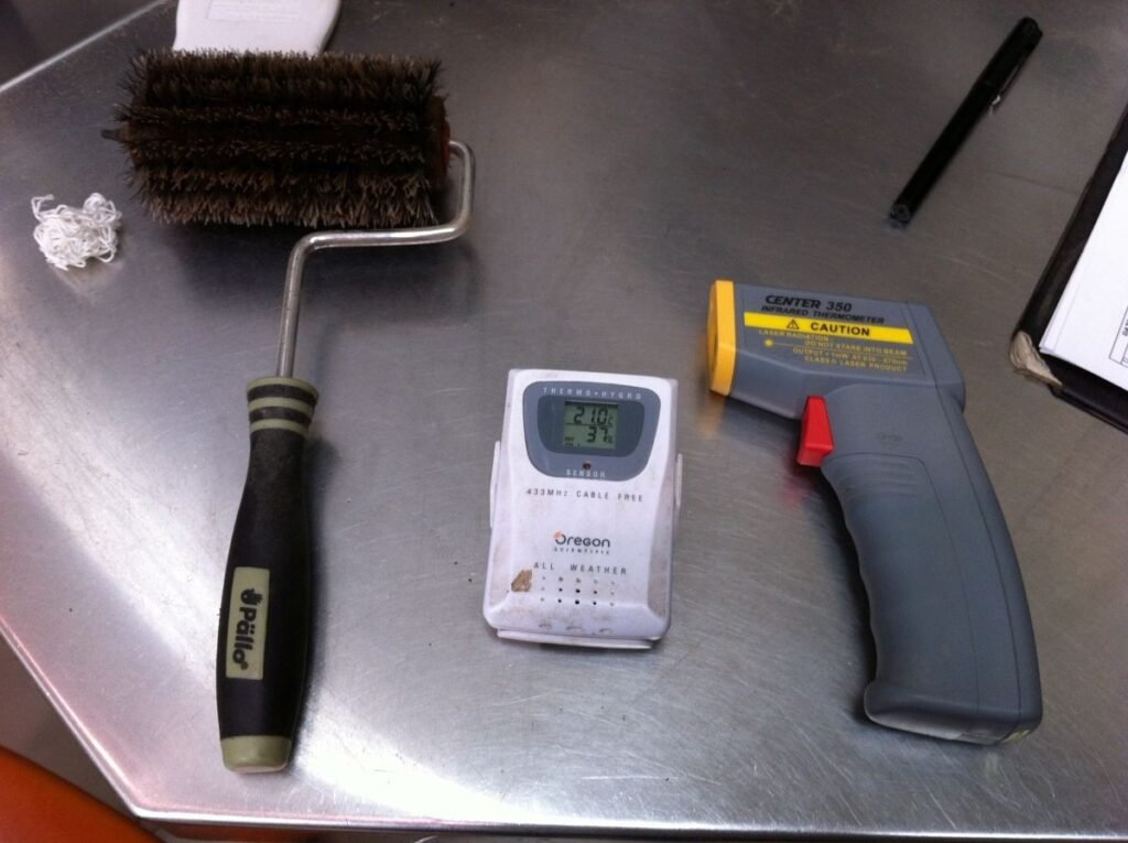 Cooling-tray brush beside a thermo-hygrometer and infrared thermometer on a stainless-steel table